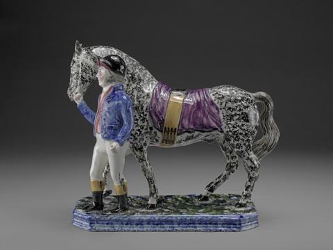Jockey and Racehorse: he in black cap, blue jacket, pink waistcoat and white breeches; the horse, sponged black with purple saddle cloth (reins missing)