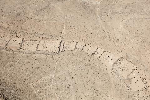 Fazal Sheikh, LATITUDE: 30°47'57"N / LONGITUDE: 34°47'3"E, October 9, 2011. Bedouin traveler (top, center right) descending a path toward the gardens near the ruined Nabataean city of Ovdat/ʽAbdāt, founded in the third century BC. During the rains, the runoff water flows downward into the streambed, which appears here in a lighter tone. The rocks that might obstruct the vegetation have been cleared away to construct low perimeter walls. The shallow light scratching marks in the image are the traces left behind by herds of sheep and goats passing through the land. Tuleilat al-ʽēnab (Arabic, “grape mounds”) can be seen on the hill in the bottom left of the image. The city was the seasonal camping ground for the Nabataean caravans traveling along the Petra–Gaza incense route., from the series Desert Bloom, 2011