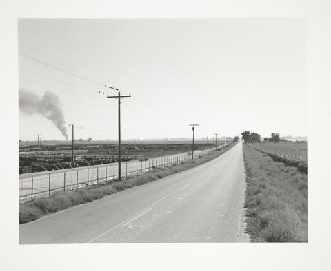 Robert Adams, Open road with smoke on horizon, feed lot, Weld County, Colorado, 1973–74, printed 2007