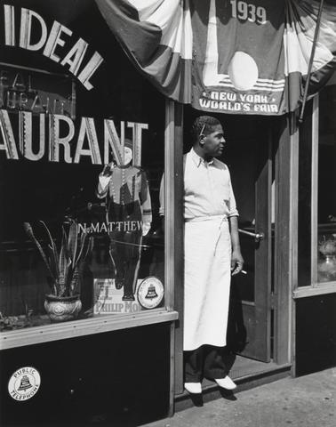 Lucien Aigner, Harlem restaurant owner standing under 1939 New York World's Fair Poster, 1939